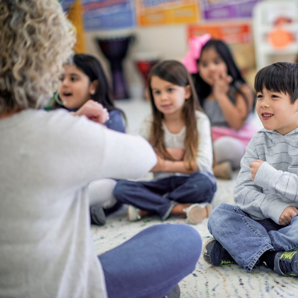 Kita Mitarbeiterin sitzt zusammen mit glücklichen Kindern auf dem Boden und singt ein Bewegungslied